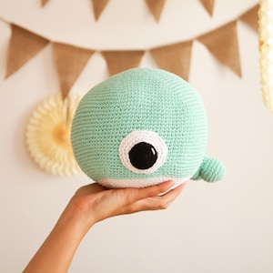 May include: A crocheted whale plushie in a soft, mint green color. The whale has a large, black eye with a white border and a small, rounded tail fin. The plushie is held in a hand, with a neutral background.