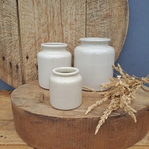 Op de afbeelding: Drie gebroken witte keramische potten van verschillende hoogtes met gladde, glanzende afwerkingen en cilindrische lichamen. De potten hebben korte, smalle halzen en brede openingen. Een klein bosje gedroogde tarwestengels ligt naast de potten.