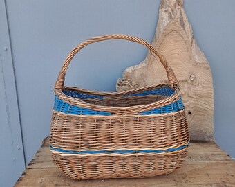 May include: A woven wicker basket featuring a natural brown finish with two horizontal bands of bright blue accent weaving. The basket has a single arched handle attached to the sides and a wide, open top with a rounded, oval-shaped base structure.