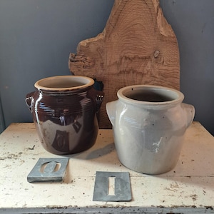 May include: Two ceramic crocks, one brown and one off-white, with side handles, sit on a distressed white table. Metal stencils with the letters "O" and "I" are in front of the crocks. A large, rough-hewn wooden slab serves as a backdrop.