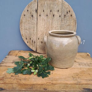 Op de afbeelding: Een beige keramische pot met twee handvatten staat naast een ronde houten snijplank met zichtbare spijkerkoppen. Een kleine tak met groene eikenbladeren en verschillende kleine groene eikels ligt voor de keramische pot.