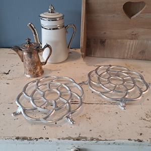May include: Two silver-colored metal trivets with a floral design are displayed on a distressed white surface. A white and silver teapot and a small silver teapot are also visible. A wooden organizer with a heart-shaped cutout is in the background.