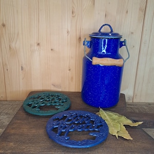 Puede incluir: Una lechera esmaltada azul con asa de madera y tapa, junto a dos salvamanteles de hierro fundido. Un salvamanteles es verde, el otro azul, ambos con la inscripción "Bon Appétit". También hay algunas hojas secas.