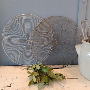 May include: Two circular, wire cooling racks with a concentric design. A bundle of green bay leaves rests on a white surface in front of the racks. A white ceramic container with kitchen utensils is on the right.