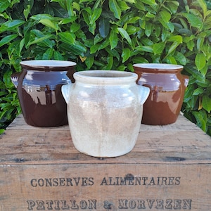 May include: Three antique ceramic jars with handles, in shades of brown and cream, sit on a wooden crate. The crate has text that reads "CONSERVES ALIMENTAIRES PETILLON & MORVEZEN QUIMPERLE FRANCE."