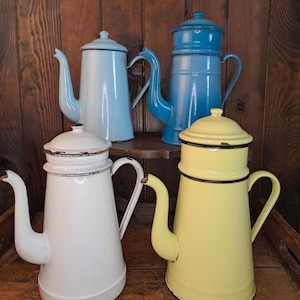 Peut inclure: Quatre cafetières anciennes en émail, dans des tons de bleu, blanc et jaune. Chaque cafetière a un bec verseur, une anse et un couvercle. Les cafetières sont exposées sur une surface en bois, mettant en valeur leur design classique et leurs couleurs.