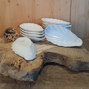 Könnte beinhalten: Eine Sammlung weißer, muschelförmiger Keramikschalen. Die Schalen sind auf einer rustikalen Holzoberfläche gestapelt und angeordnet. Ein kleiner Papierkegel mit getrockneten Blumen befindet sich ebenfalls in der Nähe. Ideal für Vorspeisen.
