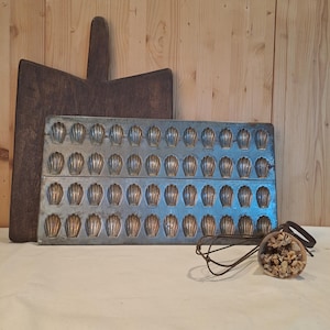 May include: A vintage baking scene featuring a dark wooden chopping board, a metal madeleine pan with shell-shaped moulds, and a wooden object with a leather strap. The items are arranged on a white surface against a wooden background.