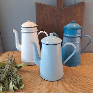 Op de afbeelding: Drie vintage emaille koffiepotten in wit en blauw. De potten hebben tuiten, handvatten en deksels. Een houten snijplank staat op de achtergrond. De potten staan op een jute oppervlak.