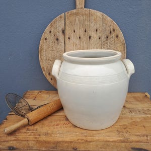 Puede incluir: Una vasija de cerámica grande de color blanco roto con dos asas laterales y detalles acanalados horizontales alrededor del cuello. El recipiente se coloca frente a una tabla de cortar redonda de madera, un rodillo de madera y una espumadera de metal.