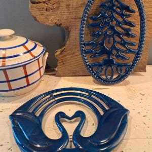 Puede incluir: Una colección de elementos decorativos azules. Incluye un salvamanteles ovalado azul con un diseño de árbol de Navidad, un salvamanteles redondo con dos figuras de cisnes y una olla de cerámica a rayas blancas y rojas con tapa.
