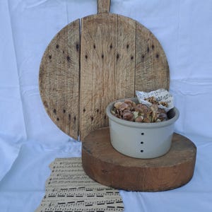 Peut inclure: Une planche à découper ronde en bois avec un manche, un bloc de bois cylindrique épais et un bol passoire en céramique gris clair rempli de pétales d'hortensia séchés et d'une partition de musique ancienne sur un tissu blanc.