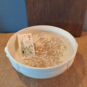 Può includere: Una ciotola in ceramica bianca con manici, riempita di fiori bianchi secchi e una cartolina vintage. La cartolina presenta un'illustrazione floreale e testo. La ciotola è appoggiata su una superficie di juta.
