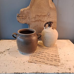 Könnte beinhalten: Ein Stillleben mit einem braunen Keramiktopf mit Henkeln, einem cremefarbenen Krug mit dunkelbraunem Griff und Noten. Im Hintergrund befindet sich eine große, unregelmäßig geformte Holzplatte. Die Objekte stehen auf einer weißen Oberfläche.