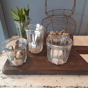 Op de afbeelding: Een rustig stilleven met heldere glazen potten en een draadmand op een houten oppervlak. Eén pot bevat groene bladeren, een andere heeft een lepel en twee andere bevatten kleine voorwerpen en kaarsen. De draadmand is gevuld met kleine eieren.