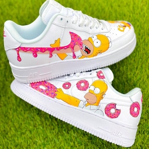 Puede incluir: Zapatillas blancas con ilustraciones personalizadas de Homer Simpson y donuts rosas. El diseño incluye a Homer Simpson comiendo una rosquilla con glaseado y chispas rosas. Los zapatos tienen la palabra "AIR" en el lateral.