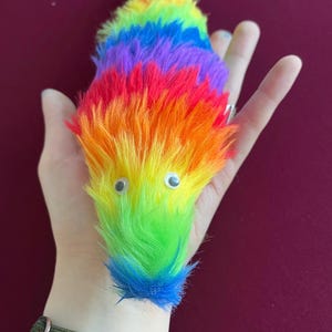 Pode incluir: Um brinquedo de verme multicolorido com olhos arregalados. O brinquedo de pelúcia apresenta um espectro de cores do arco-íris, incluindo vermelho, laranja, amarelo, verde, azul e roxo. O verme é feito de material macio e fofo.