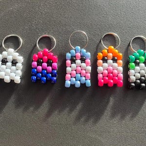 May include: Five beaded keychains in a row, each with a silver keyring. The keychains are square-shaped and feature various color combinations, including white and black, pink and blue, and orange and pink. The keychains are made of small, round beads.