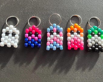May include: Five beaded keychains in a row, each with a silver keyring. The keychains are square-shaped and feature various color combinations, including white and black, pink and blue, and orange and pink. The keychains are made of small, round beads.