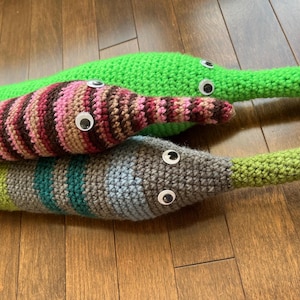 Pode incluir: Três brinquedos de pelúcia feitos à mão em crochê em forma de minhocas longas. Uma é verde brilhante sólida, uma apresenta listras horizontais rosa, marrom e bege, e a terceira tem faixas cinzas, verde-azuladas e verde-limão. Cada minhoca tem dois olhos móveis.
