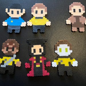 Könnte beinhalten: Pixel-Art-Figuren von Star Trek-Charakteren aus bunten Plastikperlen. Die Charaktere tragen Uniformen in Blau, Gelb und Braun. Die Figuren sind auf einer dunklen Oberfläche angeordnet.