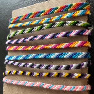 Puede incluir: Una colección de coloridas pulseras de la amistad, cada una tejida con un patrón único de hilos vibrantes. Las pulseras se exhiben en un trozo de cartón, mostrando una variedad de combinaciones de colores arcoíris, rosa, azul y negro.