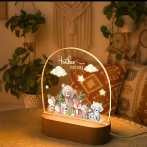 Peut inclure: Veilleuse décorative avec une scène d'animaux de la forêt, comprenant un ours, un renard, un raton laveur et un cerf. La lumière est personnalisée avec le nom "Heather" et la date 05.08.2024. La base est de couleur marron foncé.