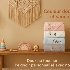 Peut inclure: Une pile de quatre serviettes à capuche pour bébés aux couleurs pastel, chacune avec un motif de visage d'animal. Les serviettes sont empilées sur une étagère en bois avec des jouets en bois et un vase avec des plantes séchées. Le texte "Léon" est visible.