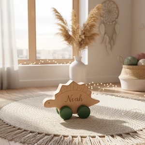 Peut inclure: Un jouet dinosaure en bois avec le nom "Noah" gravé sur le côté. Le jouet a un corps brun clair, des roues vertes et un dos épineux. Il est posé sur un tapis rond de couleur crème.