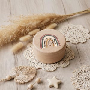 Peut inclure: Une petite boîte ronde en bois avec un motif arc-en-ciel et le nom "Lea" imprimé sur le couvercle. La boîte est posée sur un napperon en dentelle blanche, avec des éléments décoratifs.