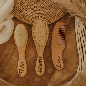 May include: Three wooden baby grooming tools: a soft brush with "Théo" engraved, an oval brush with "Maurie" engraved, and a comb with "Lisa" engraved. The tools are arranged on a wooden surface with a woven basket and dried plants in the background.