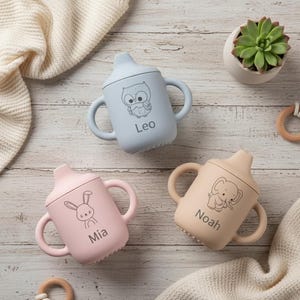 May include: Three pastel-coloured baby sippy cups. Each cup has a different animal illustration: an owl, a bunny, and an elephant. The names "Leo", "Mia", and "Noah" are printed below the illustrations. The cups are blue, pink, and beige.