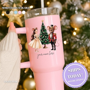 May include: Light pink tumbler with a handle, featuring a Nutcracker-themed design. The design includes a ballerina mouse, a nutcracker mouse, and a Christmas tree. The text "your name here" is also present. The tumbler has a clear lid and a straw.