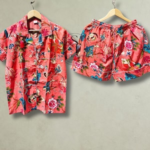Puede incluir: Un conjunto de pijama color coral que consta de una camisa de manga corta abotonada con cuello y un pantalón corto a juego. Ambas piezas presentan un estampado floral vibrante en tonos azul, blanco y verde, con ribetes blancos.