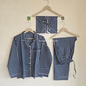 May include: Navy blue and white checkered pajama set, including a button-down shirt, pants, and a matching sleep mask, all hanging on wooden hangers. The set features white trim.