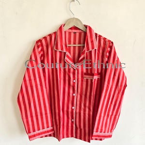 May include: Red and pink striped pajama top with white trim. The long-sleeved shirt has a button-down front, a chest pocket, and is hanging on a wooden hanger. The text "Couture Ethnic" is visible on the shirt.