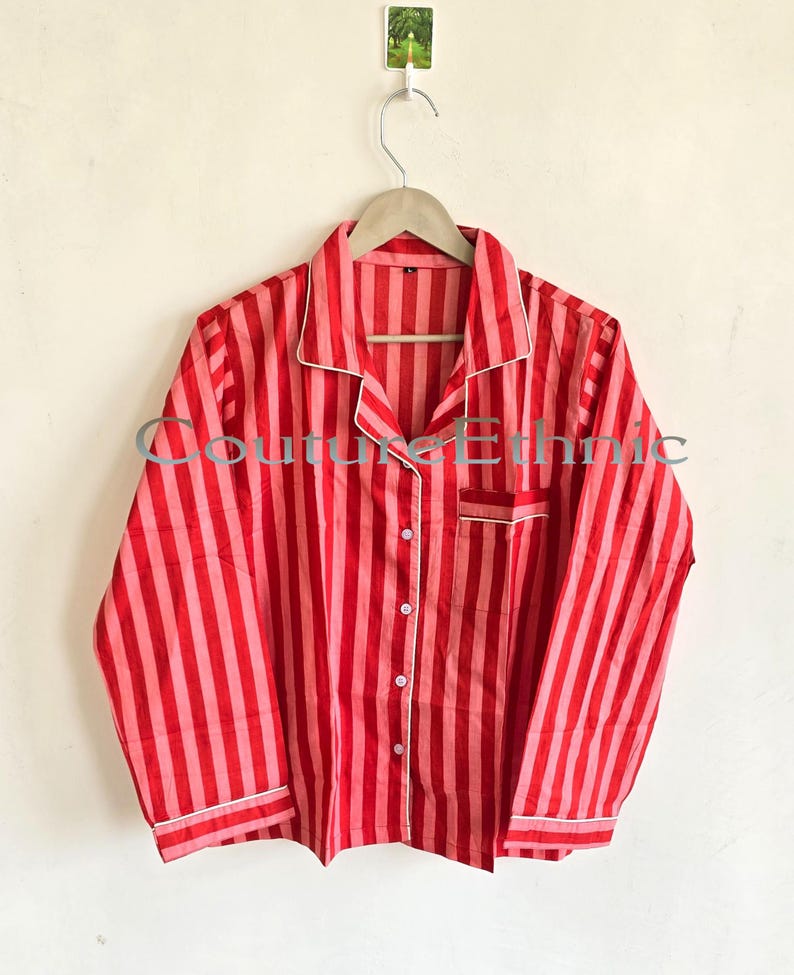 May include: Red and pink striped pajama top with white trim. The long-sleeved shirt has a button-down front, a chest pocket, and is hanging on a wooden hanger. The text "Couture Ethnic" is visible on the shirt.