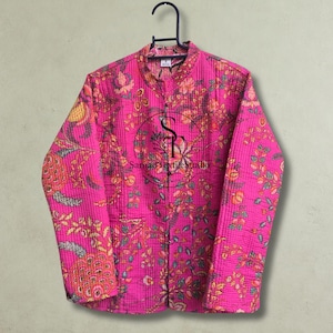 May include: A bright pink quilted jacket featuring a floral pattern in shades of orange, green, and yellow. The jacket has a mandarin collar, long sleeves, and a front button closure. A circular logo with the text Samad Textile Studio is visible.