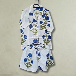 May include: A white pajama set featuring a button-down short-sleeve shirt with a collar and matching shorts. The fabric is decorated with a repeating pattern of blue flowers and green stems. The text SamadTextileStudio is visible on the shirt.