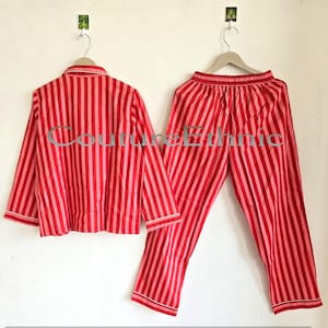 May include: A red and pink striped pajama set, including a button-down shirt and matching pants, hangs on a white wall. The shirt has a collar and long sleeves, while the pants have an elastic waistband. The set is made of a lightweight fabric.
