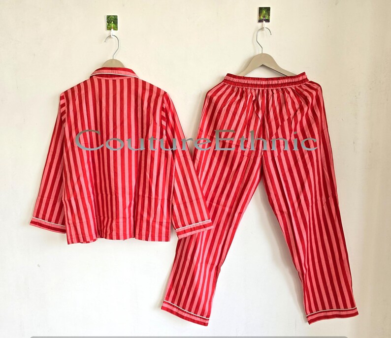 May include: A red and pink striped pajama set, including a button-down shirt and matching pants, hangs on a white wall. The shirt has a collar and long sleeves, while the pants have an elastic waistband. The set is made of a lightweight fabric.