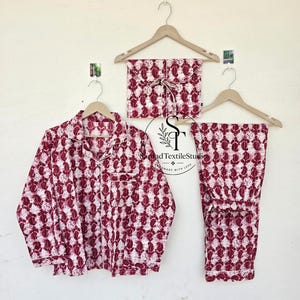 May include: A pajama set consisting of a long-sleeved button-down shirt and matching pants, featuring a dark red and white floral block print pattern. The set is displayed on wooden hangers, with a folded matching fabric pouch included above.