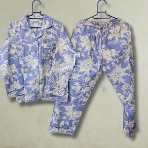 May include: A two-piece pajama set featuring a long-sleeved button-down shirt with a collar and matching pants, both in a light blue fabric with a white floral and botanical print. The shirt has dark blue piping and a chest pocket.