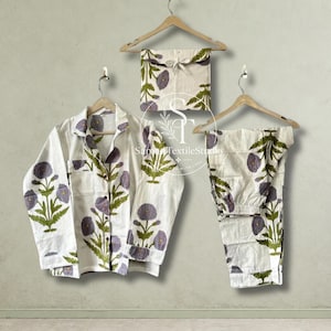 May include: A white pajama set featuring a long-sleeved button-down shirt and matching pants, both decorated with a repeating pattern of purple flowers and green stems. The set includes a folded fabric pouch with a tie closure and a logo.