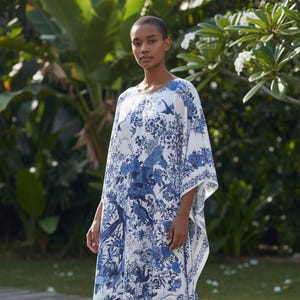 May include: A white caftan with a blue floral and bird pattern. The loose-fitting garment has a round neckline and wide sleeves. The caftan is knee-length and has side slits. The model is standing on a wooden deck in front of greenery.
