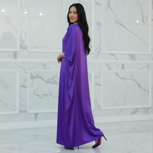 May include: A long, flowing purple dress with wide sleeves and a draped front. The dress is paired with matching purple heels. The woman is smiling and looking towards the camera. The background is a white marble wall.