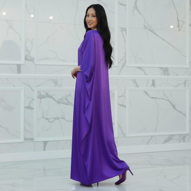 May include: A long, flowing purple dress with wide sleeves and a draped front. The dress is paired with matching purple heels. The woman is smiling and looking towards the camera. The background is a white marble wall.