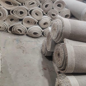 Puede incluir: Una gran colecci&oacute;n de alfombras de color blanco roto enrolladas y apiladas en un almac&eacute;n. Cada alfombra est&aacute; apretadamente enrollada, revelando el tejido texturizado del exterior y la secci&oacute;n transversal circular del material enrollado.