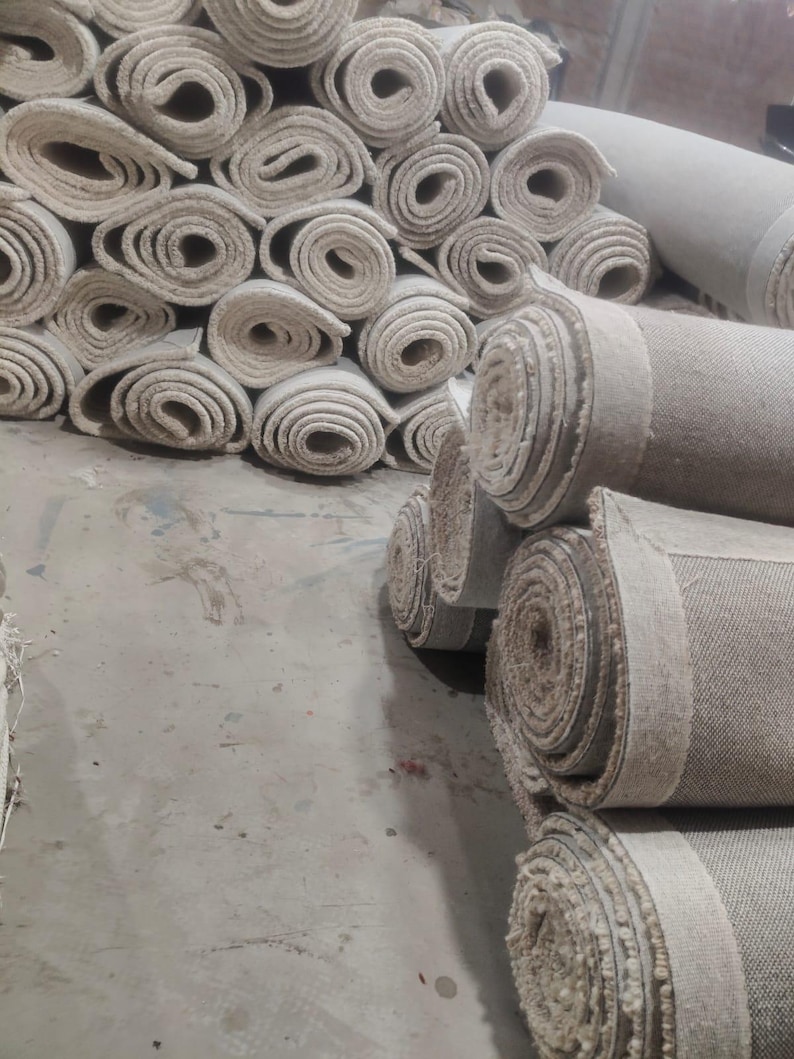 Puede incluir: Una gran colecci&oacute;n de alfombras de color blanco roto enrolladas y apiladas en un almac&eacute;n. Cada alfombra est&aacute; apretadamente enrollada, revelando el tejido texturizado del exterior y la secci&oacute;n transversal circular del material enrollado.