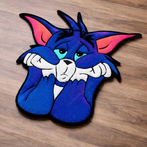Puede incluir: Alfombra azul de gato de dibujos animados con expresión triste. La alfombra presenta detalles blancos, rosas y turquesas. El gato se sujeta la cara con las patas, colocada sobre un suelo de madera.